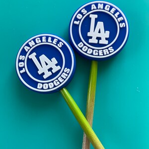 May include: Two blue and white Los Angeles Dodgers logo keychains on wooden sticks. The logos are round and feature the team's name and logo.