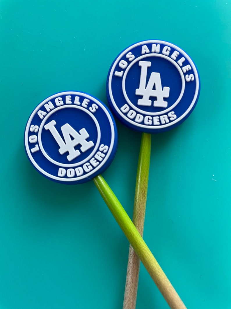 May include: Two blue and white Los Angeles Dodgers logo keychains on wooden sticks. The logos are round and feature the team's name and logo.