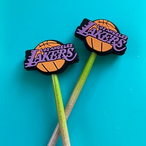 May include: Two wooden pencils with purple and gold Los Angeles Lakers basketball team logo toppers. The logos are on a black background with a basketball graphic.