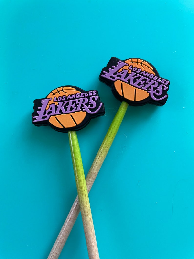 May include: Two wooden pencils with purple and gold Los Angeles Lakers basketball team logo toppers. The logos are on a black background with a basketball graphic.