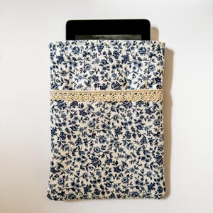 May include: A rectangular quilted fabric sleeve featuring a blue and white floral pattern. A horizontal strip of beige lace trim is stitched across the middle. A black tablet is partially inserted into the top opening of the protective case.
