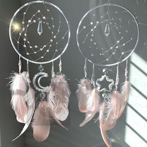 May include: Two white dreamcatchers with pink feathers and crystal accents. One dreamcatcher features a crescent moon and the other a star.