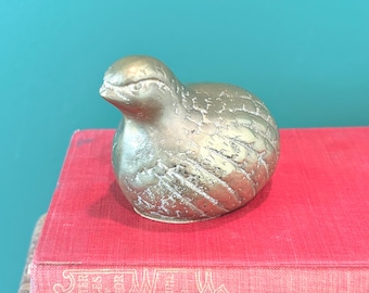 May include: A brass bird figurine with textured details, perched on a stack of red books. The bird has a rounded body and a detailed head, with a slightly weathered appearance. The background is a teal color.