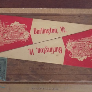 May include: Two vintage pennants with red and white designs. The pennants are on a wooden box with a stamp that says "Class C 50". The text on the pennants reads "Burlington, Vt."