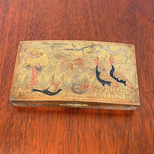 May include: A rectangular metal box featuring an intricate etched design of birds, including cranes and ducks, amidst trees and mountains. The surface displays a weathered gold-toned finish with accents of black, red, and blue paint throughout.