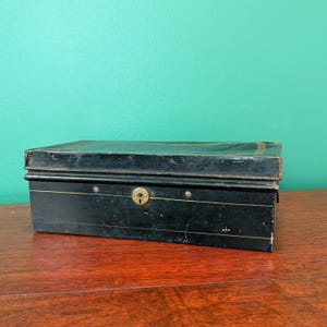 May include: A rectangular black metal box with a keyhole and brass accents. The box has a slightly distressed appearance, suggesting age. The box is sitting on a wooden surface.