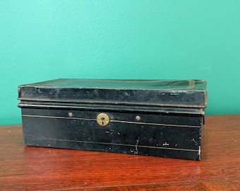 May include: A rectangular black metal box with a keyhole and brass accents. The box has a slightly distressed appearance, suggesting age. The box is sitting on a wooden surface.
