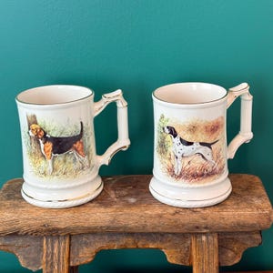 May include: Two white ceramic mugs with gold-tone rims and handles. The left mug features a tricolor hound dog in a grassy field. The right mug displays a white and black spotted pointer dog in a field. Both mugs have a rope-textured base detail.