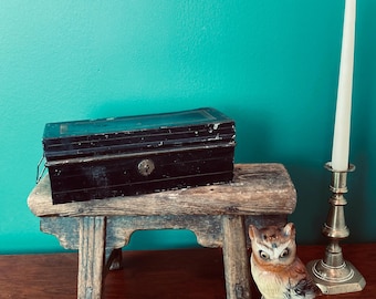 May include: A black metal box with a lock sits on a rustic wooden stool. A ceramic owl figurine and a brass candlestick with a white candle are on a wooden surface.