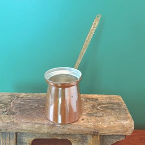 May include: A copper-colored Turkish coffee pot, also known as a cezve, with a long, ornate brass handle. The pot sits on a rustic wooden surface against a teal background. The pot's design is a classic, traditional shape.