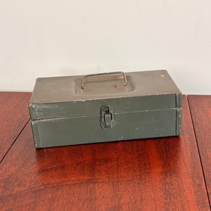 May include: A rectangular olive green metal storage box featuring a hinged lid, a central metal carrying handle on the top, and a single front-facing metal latch mechanism. The exterior surface shows signs of wear with minor paint chips and scratches.