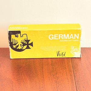 May include: A yellow box of German vocabulary cards. The box features the word "GERMAN" in white block letters, with the words "VOCABULARY CARDS" below. The box also has a black and yellow emblem and text that reads "Vis-Ed".