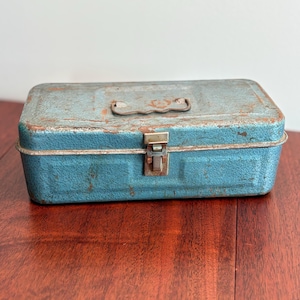 May include: A vintage, rectangular toolbox in a distressed teal finish. The metal box features a handle, a latch closure, and visible rust, suggesting age and use. The toolbox is likely used for storing tools or small items.