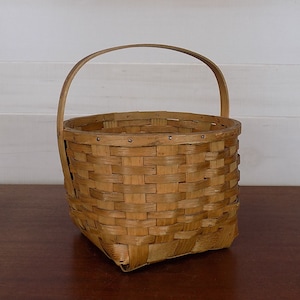 May include: A woven basket with a natural wood finish and a single handle. The basket is made of thin strips of wood that are woven together to create a sturdy and durable container.