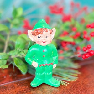 May include: A vintage ceramic elf figurine. The elf is wearing a green outfit with red accents and a matching green hat. The elf has brown hair, large ears, and a smiling face. The figurine is standing on a wooden surface, with greenery and red berries in the background.
