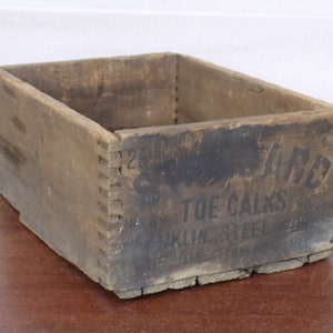 May include: A weathered wooden crate with dovetail joints. The crate has dark staining and visible text that reads "WARD TOE CALKS" and "FRANKLIN STEEL". The crate is rectangular and appears to be vintage.