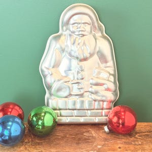 May include: A silver-colored Santa Claus-shaped mold with a detailed design, standing upright. The mold is surrounded by four colorful Christmas ornaments: red, green, and blue. The objects are on a wooden surface against a green background.