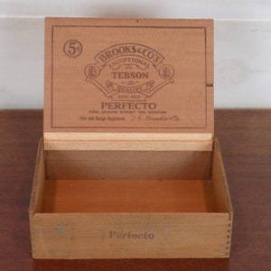 May include: A wooden box with a hinged lid. The lid has a label that reads "Brooks & Co's Exceptional Tebson Quality Very Mild Perfecto None Genuine Without The Signature Title and Design Registered T.E. Brooks & Co.". The box is labeled "Perfecto" on the bottom.