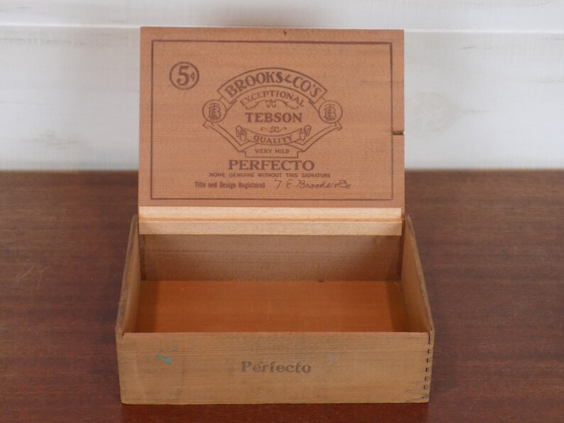 May include: A wooden box with a hinged lid. The lid has a label that reads "Brooks & Co's Exceptional Tebson Quality Very Mild Perfecto None Genuine Without The Signature Title and Design Registered T.E. Brooks & Co.". The box is labeled "Perfecto" on the bottom.