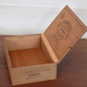 May include: A wooden box with a hinged lid. The lid is open and shows the label "Brooks & Co's Exceptional for Quality Tebson Perfecto". The box is made of light brown wood and has a natural finish.