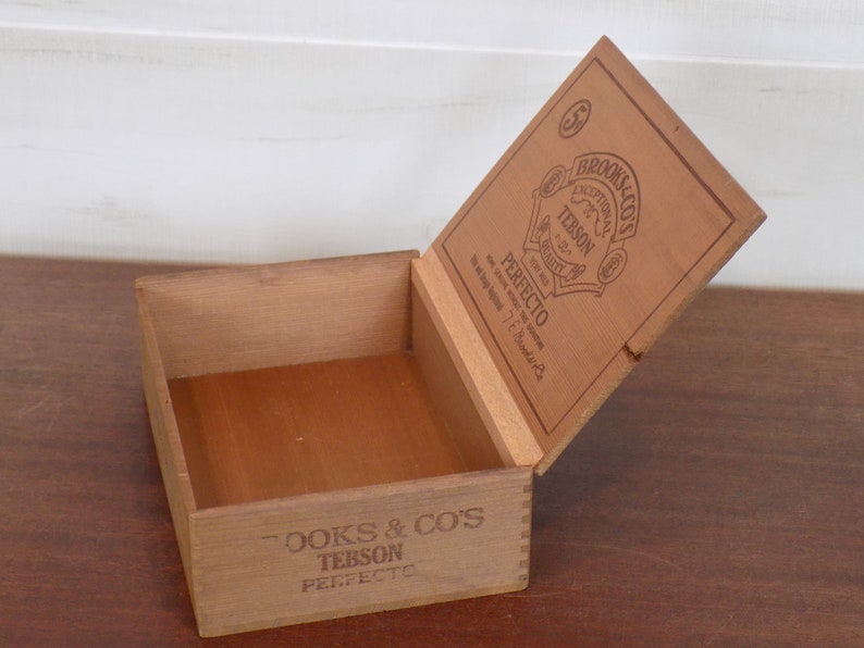 May include: A wooden box with a hinged lid. The lid is open and shows the label "Brooks & Co's Exceptional for Quality Tebson Perfecto". The box is made of light brown wood and has a natural finish.