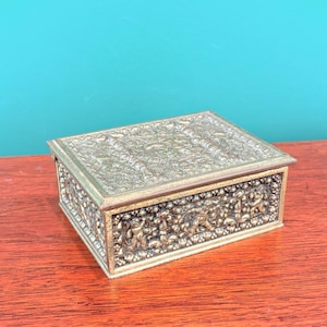 May include: A rectangular, antique brass jewelry box with intricate embossed floral and figural designs. The box has a hinged lid and is set on a wooden surface. The box is a vintage item.
