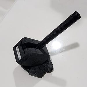 Peut inclure: Une réplique noire du marteau de Thor, Mjolnir, avec un manche texturé. La tête du marteau est gris foncé, de forme rectangulaire et présente un design détaillé. Le marteau repose sur une base rocheuse gris foncé.