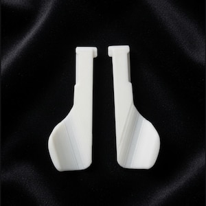 May include: Two white, L-shaped plastic components with a smooth, matte finish. Each piece features a long, straight vertical arm that transitions into a wider, curved base section. The objects are positioned side-by-side, displaying a uniform design.