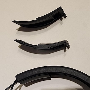 May include: Two black plastic headband replacement parts for headphones. The parts are curved and have a textured surface.
