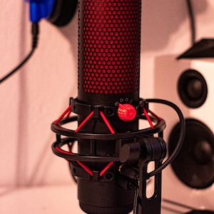 以下が含まれることがあります： 赤いメッシュグリルと赤いノブが付いた黒いマイク。マイクは、ポップフィルター付きの黒いスタンドに取り付けられています。