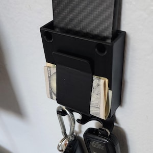 May include: A black wall mounted key holder with a carbon fiber wallet, cash, and car keys attached. The holder has a hook for keys and a slot for a wallet.