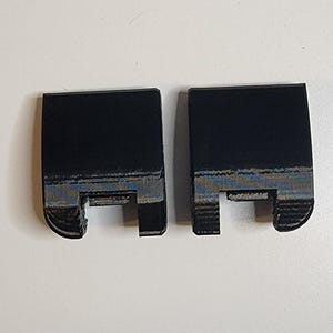 May include: Two black, rectangular plastic pieces with a notch cut out of the bottom. The pieces have a slightly textured surface and are positioned side-by-side.