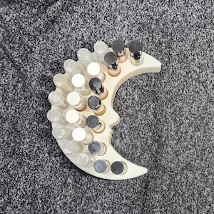 May include: A white, crescent moon-shaped organizer holds small glass bottles with various colored contents. The organizer has a profile cutout and is on a textured surface. The bottles have white, black, and silver caps.