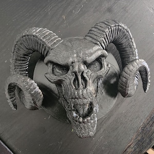 May include: A black and gray skull with horns, the skull has a detailed textured surface and an open mouth with teeth. The skull is mounted on a black circular base.
