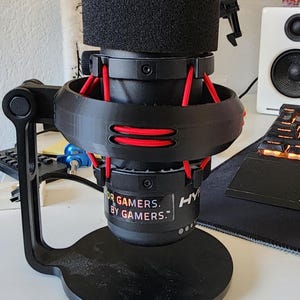 May include: A black microphone with red detailing, mounted on a black stand. The microphone features a black foam cover and the text "FOR GAMERS. BY GAMERS." is printed on the side.