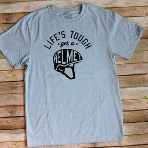 Op de afbeelding: Een lichtblauw t-shirt met zwarte tekst die "Life's Tough, get a Helmet" luidt en een zwart silhouet van een helm.