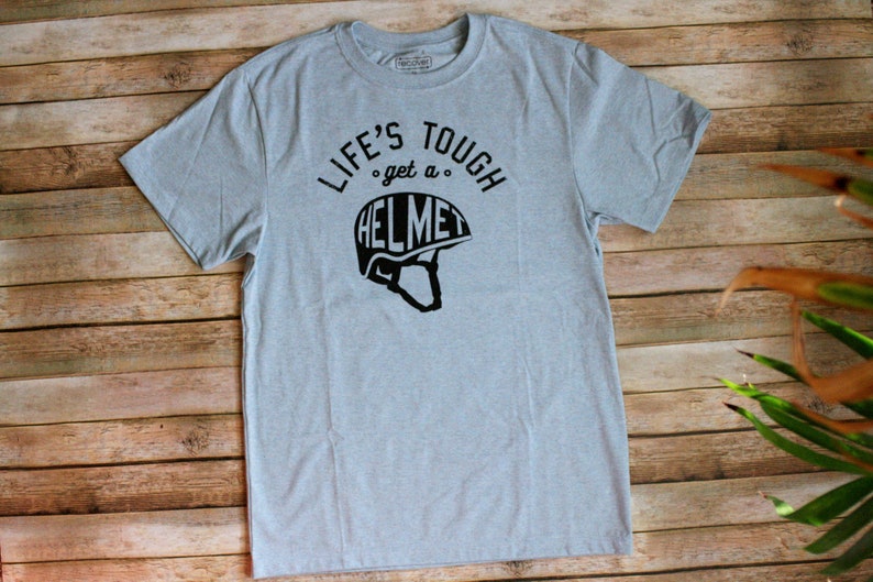 Op de afbeelding: Een lichtblauw t-shirt met zwarte tekst die "Life's Tough, get a Helmet" luidt en een zwart silhouet van een helm.