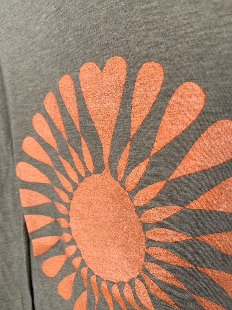 May include: A gray t-shirt with a large orange sun graphic. The sun is made up of a circle with radiating lines that resemble petals.