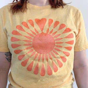 May include: A yellow t-shirt with a large orange sun graphic. The sun has a heart shape in the center and is surrounded by rays.