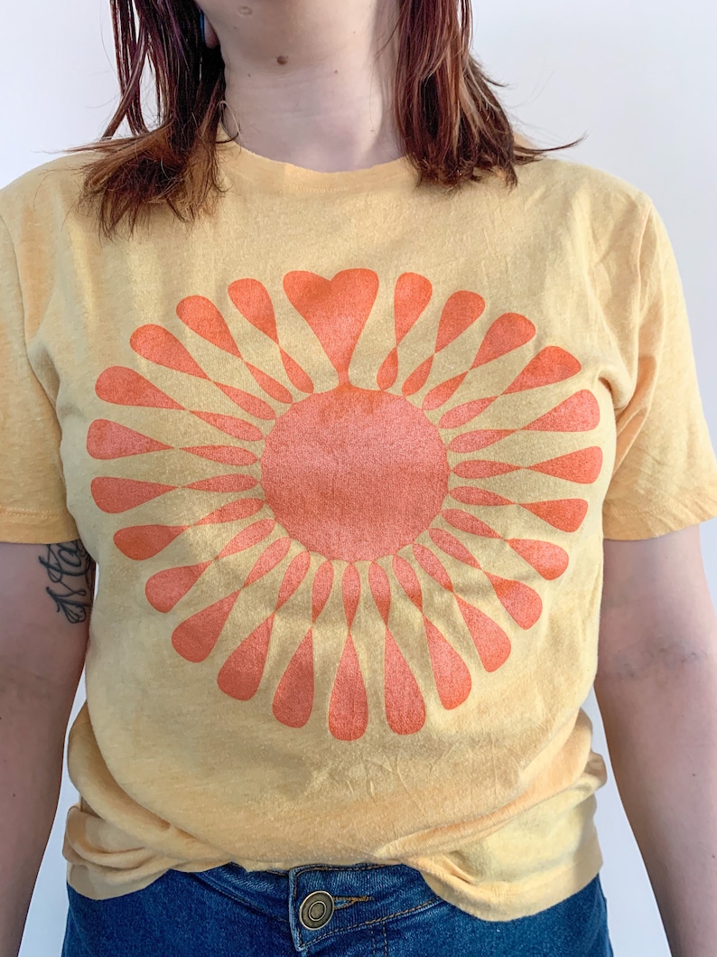 May include: A yellow t-shirt with a large orange sun graphic. The sun has a heart shape in the center and is surrounded by rays.