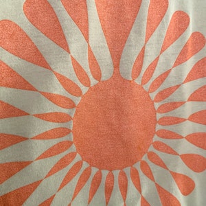 May include: A white t-shirt with a large, peach-colored sun design. The sun is made up of a circle with radiating rays that are shaped like teardrops.