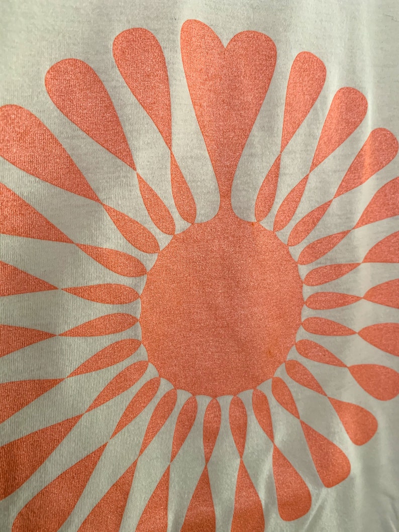 May include: A white t-shirt with a large, peach-colored sun design. The sun is made up of a circle with radiating rays that are shaped like teardrops.