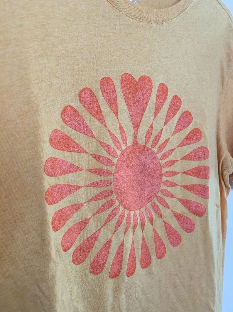 May include: A yellow t-shirt with a pink sunburst design. The sunburst is made up of a circle of pink hearts.
