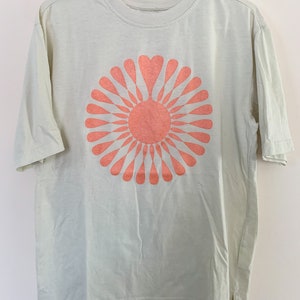 May include: A light green t-shirt with a peach-colored sunburst design. The sunburst has a heart shape in the center.
