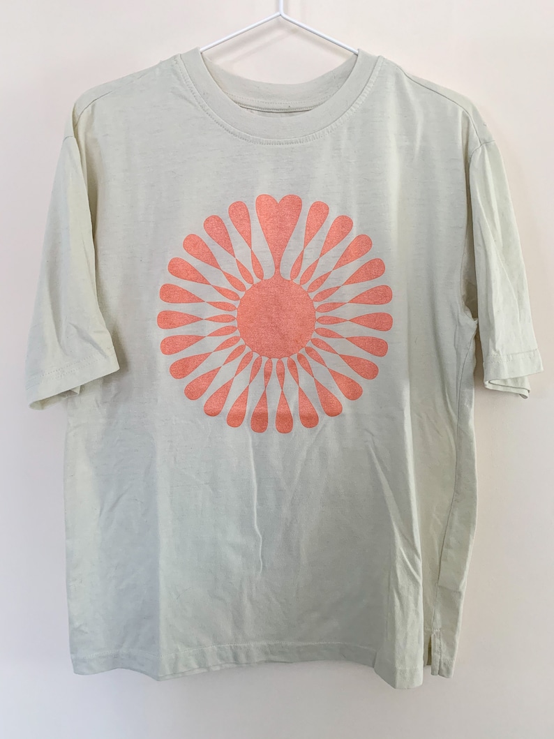 May include: A light green t-shirt with a peach-colored sunburst design. The sunburst has a heart shape in the center.