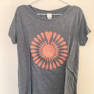 May include: A gray t-shirt with a peach-colored graphic of a stylized sun with heart-shaped petals.