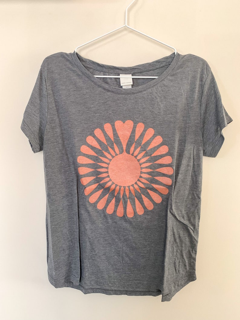 May include: A gray t-shirt with a peach-colored graphic of a stylized sun with heart-shaped petals.