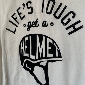 Op de afbeelding: Wit T-shirt met zwarte tekst die "Life's Tough, get a Helmet" luidt. De tekst is cirkelvormig met een helm-afbeelding eronder.