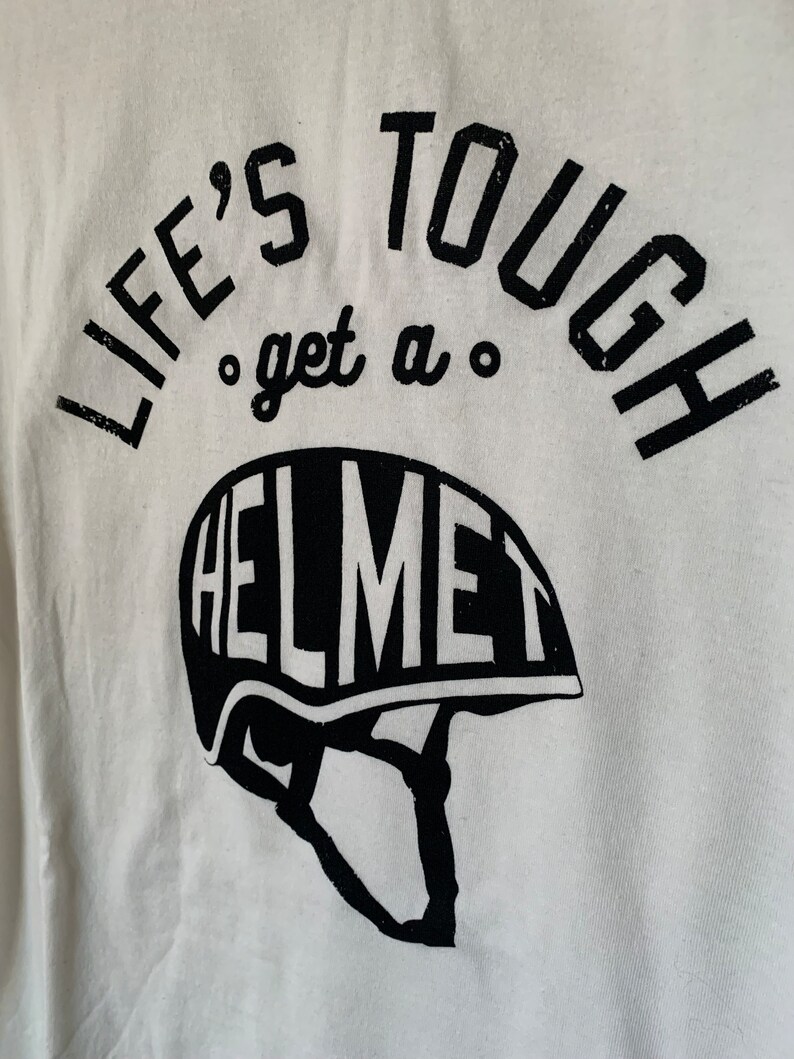 Op de afbeelding: Wit T-shirt met zwarte tekst die "Life's Tough, get a Helmet" luidt. De tekst is cirkelvormig met een helm-afbeelding eronder.