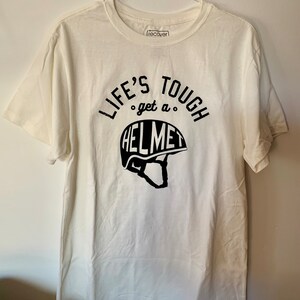 Op de afbeelding: Wit T-shirt met zwarte tekst die "Life's Tough get a HELMET" zegt met een afbeelding van een helm.